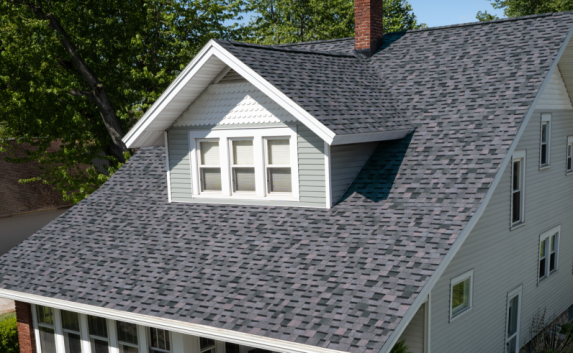 Shingle color of the year newly installed on the roof of a small gray home with slanted roof and one large dormer in the middle. Trees in background.