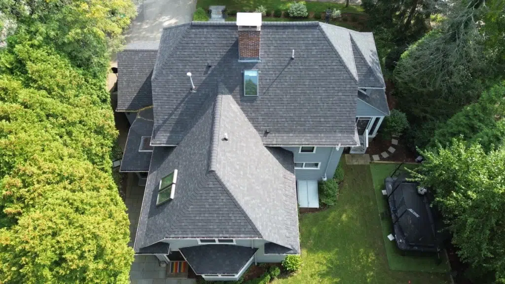 Aerial view of a large residential home with gray shingle roofing surrounded by lush greenery and a well-manicured lawn.